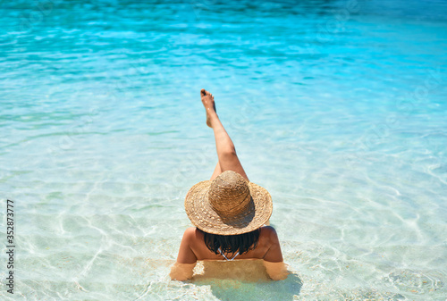 Back view of young woman in straw hat and white bikini relax in turquoise sea on tropical paradise beach