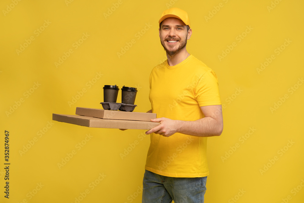 Portrait of young delivery man in yellow uniform with pizza boxes and takeaway coffee isolated over yellow background. Portrait of young delivery man in yellow uniform with pizza boxes and takeaway coffee isolated over yellow background.
