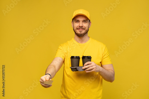 Portrait of young delivery man in yellow uniform with takeaway coffee isolated over yellow background.