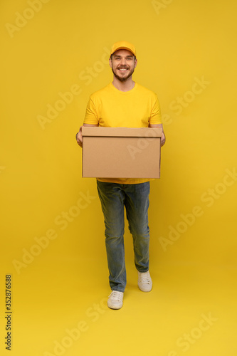 Portrait of excited delivery man in yellow uniform holding paper box isolated over yellow background.