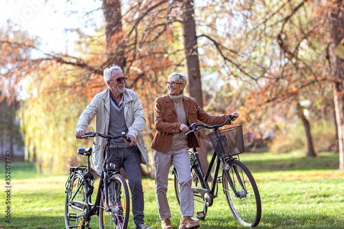 Mature fit couple pushing bicycles in public park talk and smile