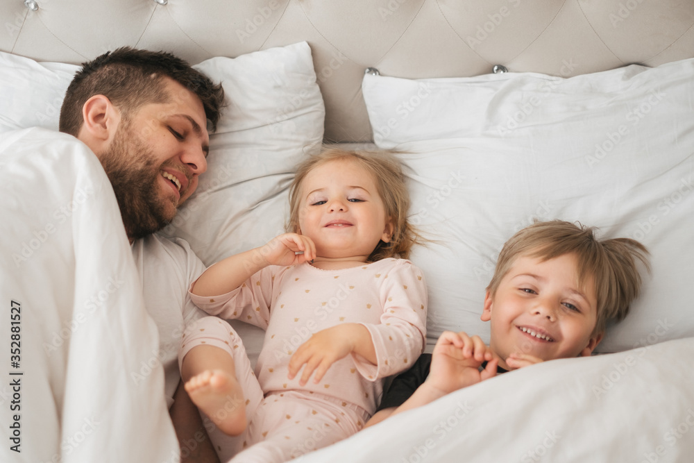 Young caucasian father plays with children in bed weekend. Cuddle daughter and son in the morning. Close up family portrait. Father's day concept