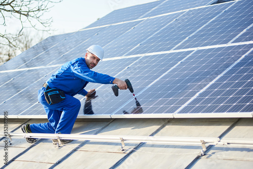 Male worker in blue suit and protective helmet installing solar panel system using screwdriver. Electrician mounting solar module on roof of modern house. Alternative energy ecological concept.