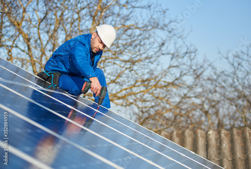 Male worker in blue suit and protective helmet installing solar photovoltaic panel system using screwdriver. Electrician mounting blue solar module on roof of modern house. Alternative energy concept.