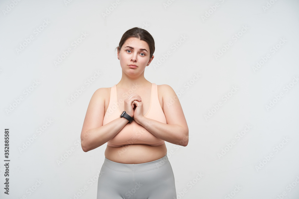Upset young pretty dark haired oversized female folding hands on her chest and looking at camera with upset face, standing over white background in nude top and leggins Upset young pretty dark haired oversized female folding hands on her chest and looking at camera with upset face, standing over white background in nude top and leggins