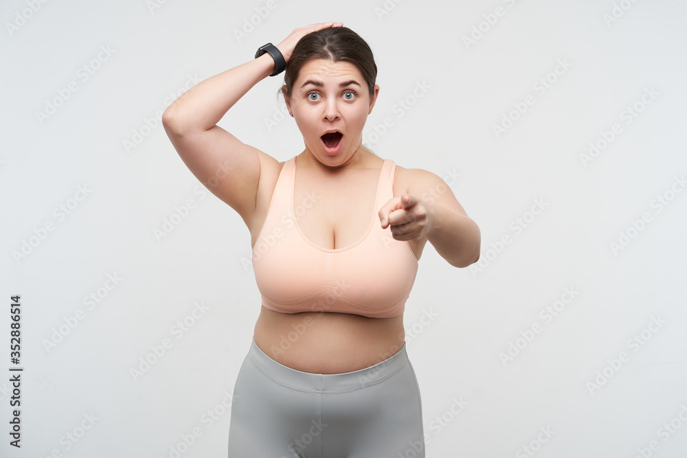 Indoor photo of young lovely brunette oversized woman in sports bra and leggins pointing dazedly at camera with wide mouth opened, isolated over white background Indoor photo of young lovely brunette oversized woman in sports bra and leggins pointing dazedly at camera with wide mouth opened, isolated over white background