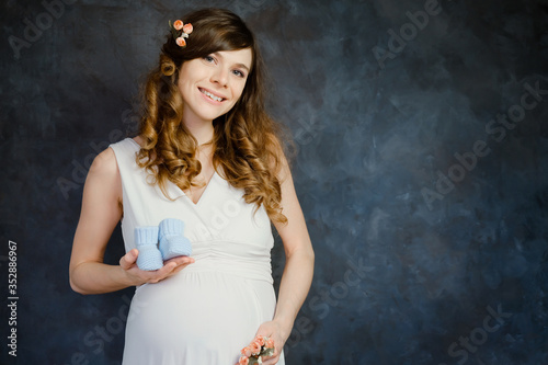 Pregnant girl with baby booties smiling standing over dark backdrop. Pregnancy, maternity concept.