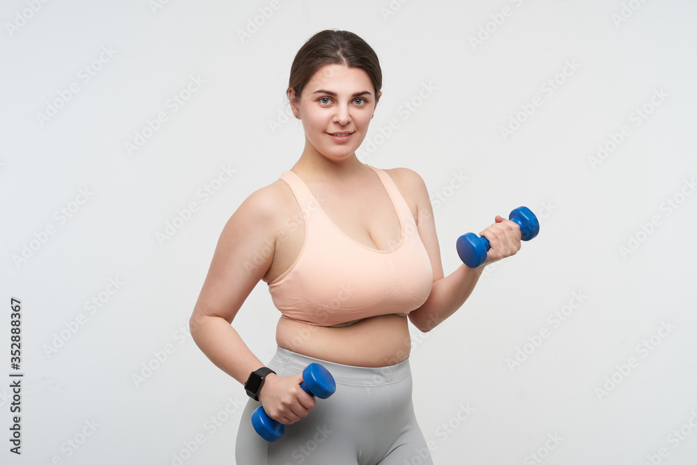 Studio photo of young positive oversized female exercising with dumbbells and looking gladly at camera while posing over white background. Concept of weight loss Studio photo of young positive oversized female exercising with dumbbells and looking gladly at camera while posing over white background. Concept of weight loss