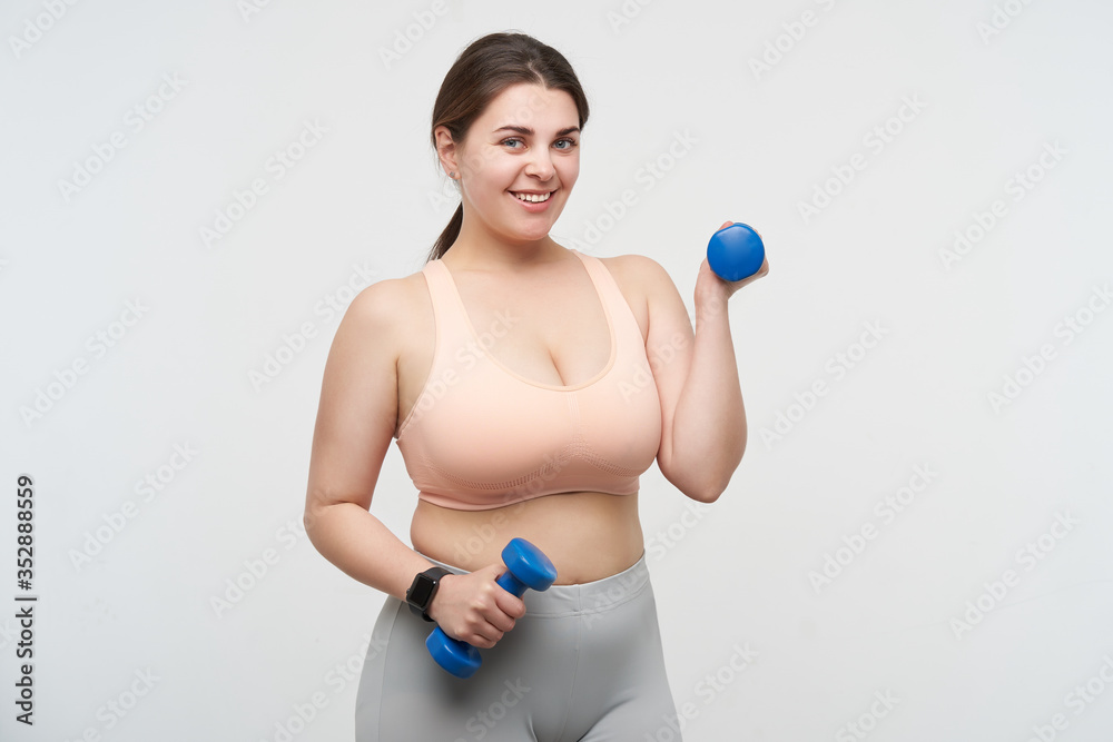 Positive young plus size model with ponytail hairstyle making sports while posing over white background and smiling gladly at camera. Body positive concept Positive young plus size model with ponytail hairstyle making sports while posing over white background and smiling gladly at camera. Body positive concept