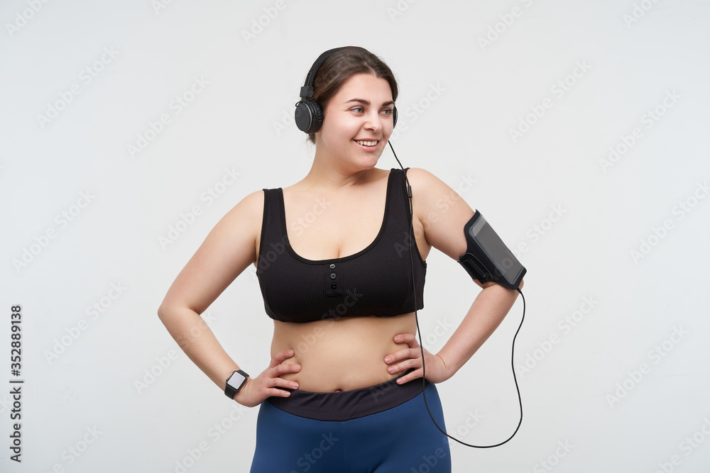 Glad young lovely dark haired fatty female with casual hairstyle holding hands on her waist and looking aside with wide smile while posing with headphones over white background Glad young lovely dark haired fatty female with casual hairstyle holding hands on her waist and looking aside with wide smile while posing with headphones over white background