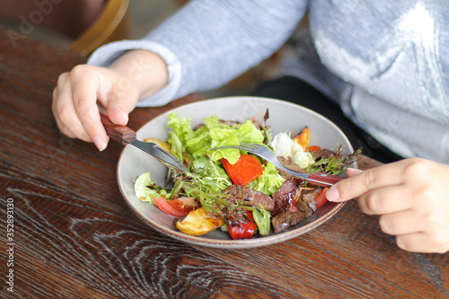 Woman is eating healthy vegetables and beef salad with knife and fork on wooden table