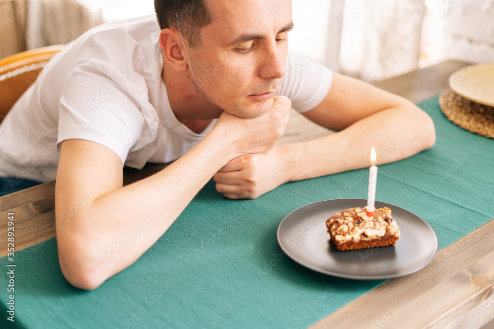 Upset young man sitting at the birthday cake and looking with sad eyes on it. Concept of celebrating alone.