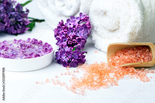 Sea salt, rolled white towels and lilac blossom flowers on white background. Spa products, skin and body care concept. 