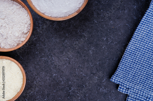 Dark marble background with rice flour, corn flour and whole grain flour in wooden bowls and blue napkin. Baking ingredients with space for text or image. Flat lay top view, studio shot