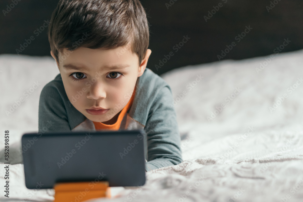 selective focus of adorable, focused boy watching online lesson on smartphone in bedroom selective focus of adorable, focused boy watching online lesson on smartphone in bedroom