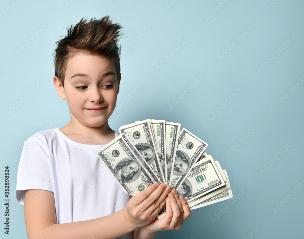 Stylish kid looking on fan of dollar banknotes in his hands with admire imagining how much he can purchase. Half-length portrait isolated on blue Stylish kid looking on fan of dollar banknotes in his hands with admire imagining how much he can purchase. Half-length portrait isolated on blue