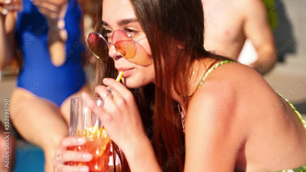 Pretty mixed race woman in stylish sunglasses, swimsuit chilling on pink inflatable flamingo, drinking cocktail of straw at pool party. Friends having fun sunbathing with drinks sitting at poolside. Pretty mixed race woman in stylish sunglasses, swimsuit chilling on pink inflatable flamingo, drinking cocktail of straw at pool party. Friends having fun sunbathing with drinks sitting at poolside.