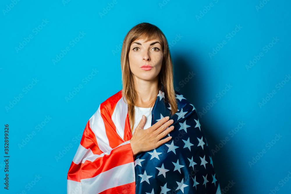 A young woman wrapped in the USA flag makes a hand gesture on a blue background. USA visa concept, English language, gesture is all right, thumbs up, like