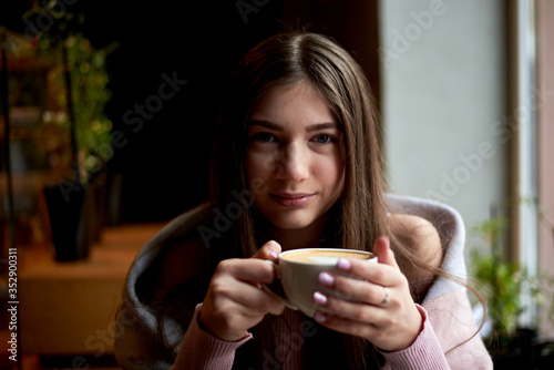 Young brunette girl, wearing pink jumper, drinking cappuccino in cozy cafe with green plants by the window in winter spring autumn, thinking. Student having lunch break in coffee house. Sunday leisure