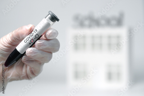 a doctor’s hand in a glove holds a test tube with a vaccine, against the background of the school
