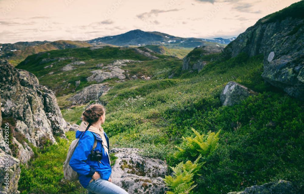 Naklejka premium Young woman traveler in a blue jacket with a backpack and a photo camera in the mountains, against the backdrop of a beautiful landscape