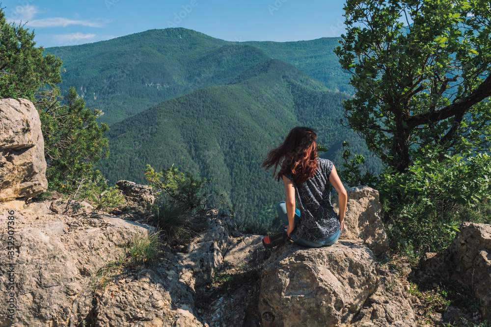 A full body shot of an unrecognizable young Caucasian female hiker sitting on a rock in the French Alps mountains on a sunny summer day