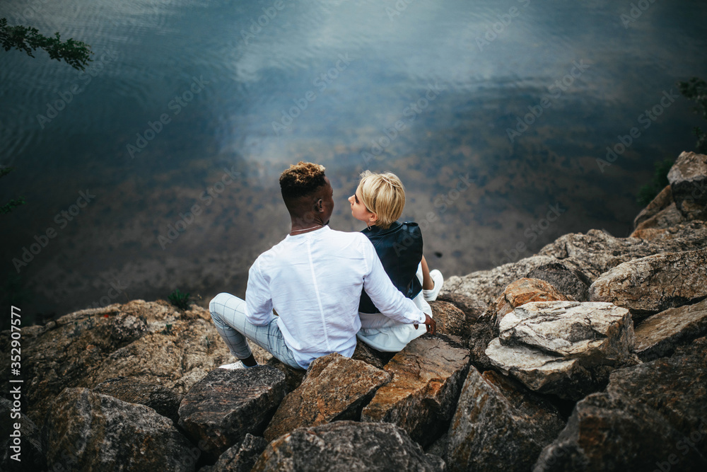 Interracial couple sits on rocks and hugs against background of river.