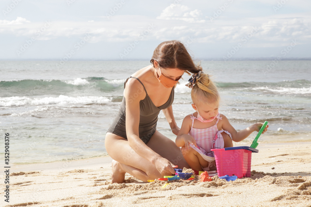 Summer vacation after Coronavirus pandemic crisis. Adorable toddler girl and her mum wearing protective mask while playing with beach toys on the sandy beach. - obrazy, fototapety, plakaty 