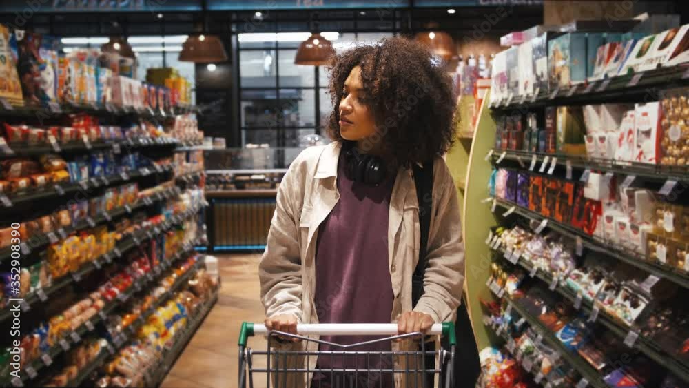 Ethnic black girl in casual clothes. She walking with shopping cart along counter with biscuits and candies and choosing sweets from grocery. Close up