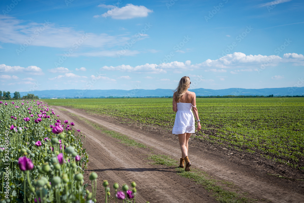 Adult caucasian woman in white dress walking through agricultural fields on sunny day with purple flowers and perfect blue sky