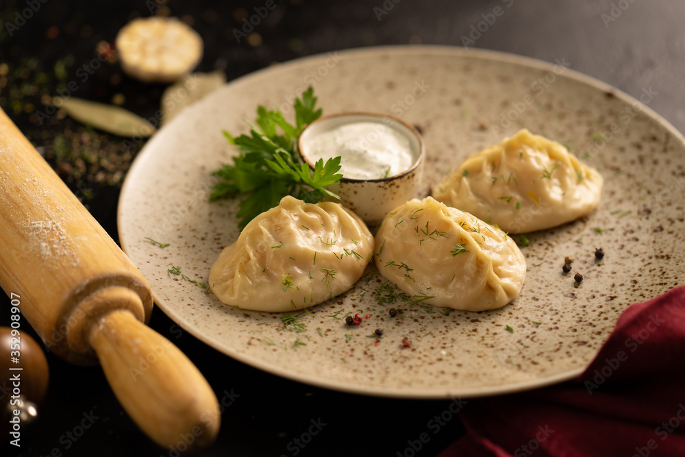 Homemade khinkali with meat, dill, sour cream sauce, garlic and bay leaf near a rolling pin on a black table. Blurred background. Homemade khinkali with meat, dill, sour cream sauce, garlic and bay leaf near a rolling pin on a black table. Blurred background.