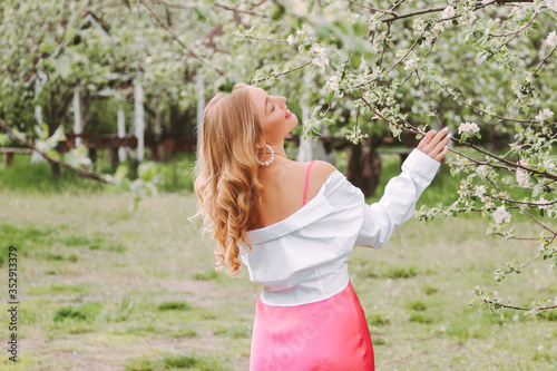 Back view stylish young beautiful hippie girl in white shirt and pink dress relax and enjoy summer nature on green garden. Happy attractive blonde woman posing with blossom tree in countryside park