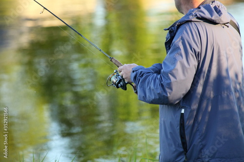 A fisherman in a jacket with a spinning rod in his hand winds a fishing line on a reel close-up on blurred water with shore trees reflection background, fishing on the river on a summer day © Ilya