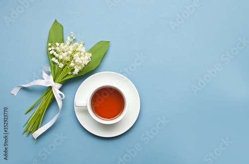 Lily of the valley flowers and cup of tea on blue background. Flat lay, top view, copy space.