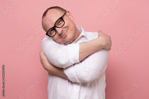 Mature caucasian man hugging himself against pink background. Positive facial human emotion,