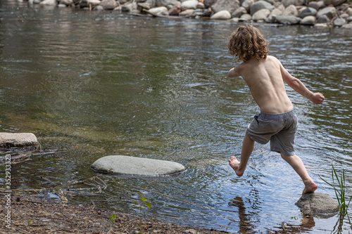 A young boy without his shirt is jumping over rocks in a pond. He is playing in the water, in the summer at camp. Leaping across stones in the creek, pond, lake. Summer vacation, camp. Fun in sun