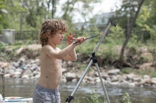 A boy is painting outside in the woods, the kid is shirtless in the summer. He is creative, having fun with art, paint and his imagination in nature. He is by a lake, pond, stream of water. 