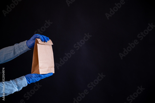 Female hands in a blue glove holds a brown paper bag on a black background. Safe food delivery to your home. A courier in a denim shirt holds out a craft cardboard bag to a customer.