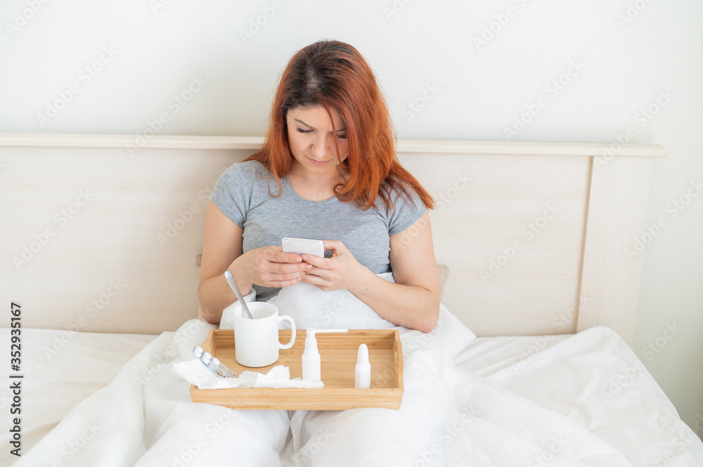 Caucasian woman on sick leave is sitting in bed with drugs on a wooden tray and is typing in the phone. A red-haired girl with a cold lies at home and uses a smartphone. Caucasian woman on sick leave is sitting in bed with drugs on a wooden tray and is typing in the phone. A red-haired girl with a cold lies at home and uses a smartphone.