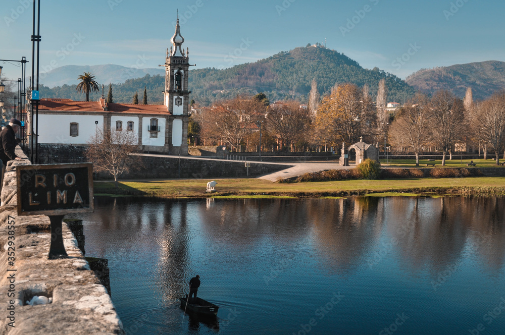 Church on the river bank. The boat floats along the river with her husband inside. In the background mountains and park. Ponte de Lima  Portugal.