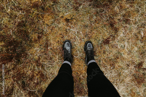 Looking down on female legs wearing black boots and leggings standing on yellow grass field