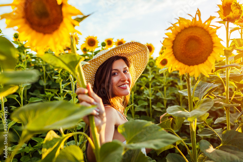 Young happy woman walking in blooming sunflower field wearing hat feeling free and admiring landscape. Summer vacation