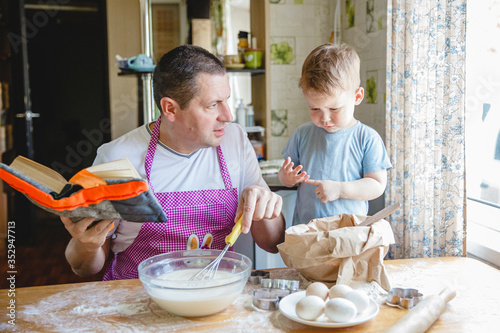 A father with a young son at the kitchen table preparing dough. Dad is reading a book, and the boy is busy in the flour.