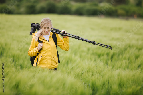 Beautiful mature woman in yellow jacket and black trousers walking among green wheat field and carrying on professional tripod for photo shoot. Concept of favorite hobby and nature beauty