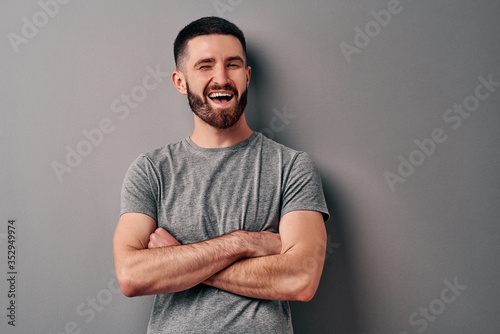 Quadro em tela Portrait of a handsome young man leaning against a gray wall crossed his arms and winks and smiles