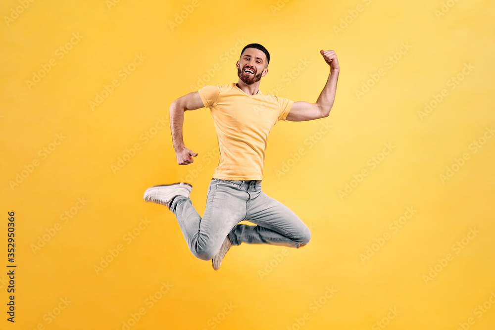 Let's dance in the jump! Young man wearing a yellow t-shirt and light jeans jumping against a background of yellow wall showing strong hands.
