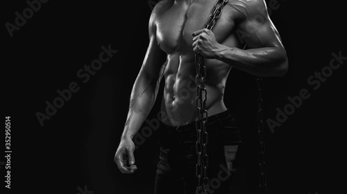 Muscular model sports young man on dark background. Black and white fashion portrait of strong brutal guy with chain. Sexy torso. Male flexing his muscles. Sport workout bodybuilding concept.