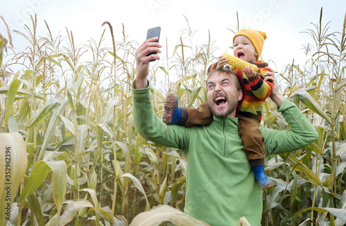 Father carries little son in his shoulders and make selfie while walking across yellow autumn corn field. Fall season concept.