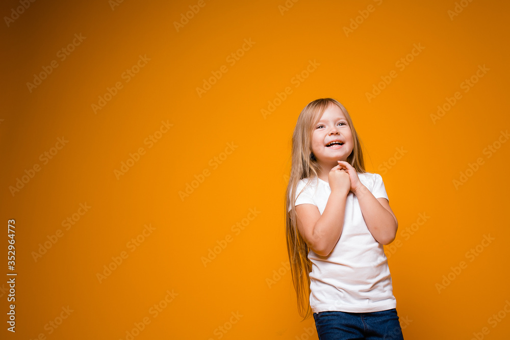 Long-haired little girl holds her hands near her chest and smiles happily in anticipation of a gift on an orange background Long-haired little girl holds her hands near her chest and smiles happily in anticipation of a gift on an orange background
