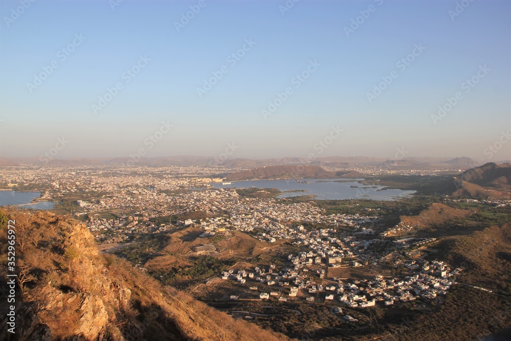 Panorama View of the City of Udaipur in Rajasthan, India, at Cloudless Sunset with a Dusty Yellow Background Hue and a Sprawl of Mostly White Houses in the Foreground